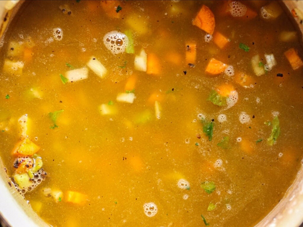 A close-up of vegetable soup simmering, showing chunks of carrots, celery, and onions in a golden broth with small bubbles and specks of herbs floating on the surface.